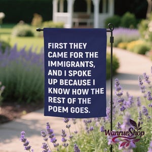 May include: A navy blue garden flag with white text that reads "FIRST THEY CAME FOR THE IMMIGRANTS, AND I SPOKE UP BECAUSE I KNOW HOW THE REST OF THE POEM GOES." The flag is displayed in a garden with lavender plants and a white gazebo in the background.