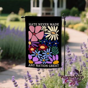 May include: Black garden flag featuring the text "HATE NEVER MADE ANY NATION GREAT" above a vibrant floral arrangement. The design includes pink, white, blue, and purple flowers. The flag is displayed in a garden with lavender plants.