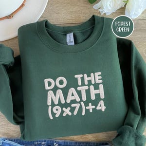 May include: Forest green sweatshirt with the text "DO THE MATH (9x7)+4" in white. The sweatshirt is folded, with a white hat and a flower in the background. The sweatshirt is made of a soft material.