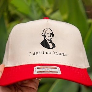May include: A beige and red baseball cap with the text "I said no kings" and a black silhouette of George Washington. The cap has a red brim and a red button on top.