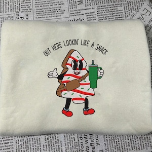 May include: Cream-colored sweatshirt with an embroidered cartoon gingerbread man holding a green tumbler. The gingerbread man has a brown body, red shoes, and a brown satchel. Text above reads "OUT HERE LOOKIN' LIKE A SNACK."