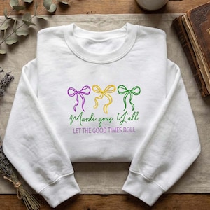 Puede incluir: Sudadera blanca con lazos bordados morados, amarillos y verdes. El texto "Mardi Gras Y'all" y "Let the Good Times Roll" están debajo. Una prenda de estilo casual.