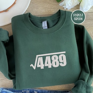 May include: Forest green sweatshirt with the square root of 4489 in white. The sweatshirt is folded on a wooden surface. A white hat and a flower are in the background. The text "Forest Green" is in a white circle.
