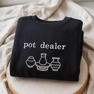 May include: Black sweatshirt with the text "pot dealer" in white, above three white line-drawn pottery designs. The sweatshirt is displayed on a neutral-colored surface.