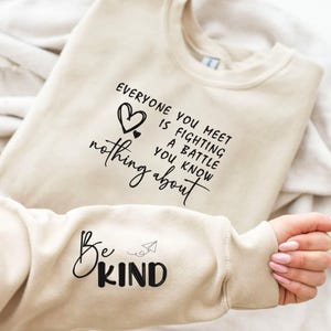 Może przedstawiać: Beżowa bluza z czarnym tekstem. Przód bluzy głosi "EVERYONE YOU MEET IS FIGHTING A BATTLE YOU KNOW NOTHING ABOUT" z sercem. Rękaw głosi "Be KIND" z papierowym samolotem.