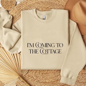May include: A beige crewneck sweatshirt with the text "I'M COMING TO THE COTTAGE" in black. The sweatshirt is displayed on a woven surface with a hat and a dried palm leaf.