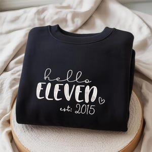 May include: Black crew neck sweatshirt featuring the text "hello ELEVEN est: 2015" in white script and block letters. A small white heart is next to the word "ELEVEN". The sweatshirt is folded on a neutral-coloured surface.