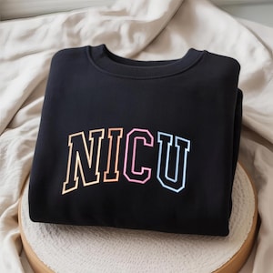 May include: Black crewneck sweatshirt with the word "NICU" in a multi-colored, collegiate-style font. The letters are outlined in white and filled with pastel colors. The sweatshirt is folded neatly on a light-colored surface.