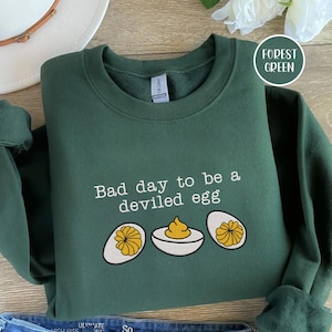 Könnte beinhalten: Forstgrünes Sweatshirt mit dem Schriftzug "Bad day to be a deviled egg" und einer Grafik von gefüllten Eiern. Das Sweatshirt ist auf einer Holzoberfläche gefaltet. Das Gildan-Markenschild ist sichtbar.