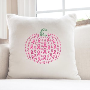May include: A white square pillow with a pumpkin design made of pink ribbons. The pumpkin stem is made of green ribbons. The pillow is on a light-colored sofa near a window.