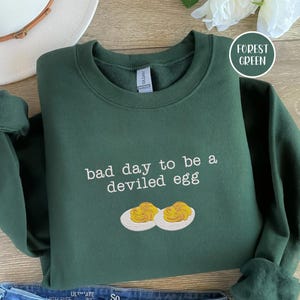 May include: A forest green sweatshirt featuring the text "bad day to be a deviled egg" and an embroidered image of two deviled eggs. The sweatshirt is folded on a wooden surface. The colour is described as "Forest Green".