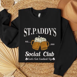 Puede incluir: Sudadera negra con el texto blanco "ST. PADDY'S Social Club", con dos jarras de cerveza con espuma. También está presente el texto "EST 2026". Debajo, "Let's Get Lucked Up" con gráficos de tréboles.