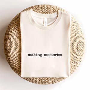 Pode incluir: Camiseta creme com a frase "making memories." impressa em preto no centro. A camiseta está dobrada e colocada sobre uma superfície circular tecida. A camiseta é feita de um material macio.