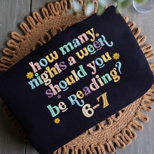 May include: Black zippered pouch with embroidered text in pastel colors: "How many nights a week should you be reading? 6-7." The pouch is on a brown woven placemat.