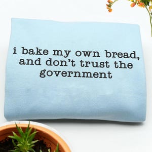 Könnte beinhalten: Ein hellblaues, gefaltetes Handtuch mit dem schwarzen Text: "i bake my own bread, and don't trust the government". Das Handtuch liegt auf einer weißen Oberfläche, mit einer Topfpflanze und Blumen im Hintergrund.