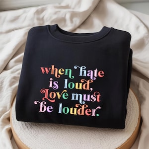 Può includere: Felpa nera con la scritta ricamata "when hate is loud, love must be louder" nei colori dell'arcobaleno. La felpa è piegata e appoggiata su una superficie dai toni neutri.