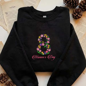 Puede incluir: Sudadera negra con un diseño bordado floral del número ocho en rosa, amarillo, morado y verde. El texto "Women's Day" está bordado en rosa debajo del número. La sudadera se muestra sobre una superficie con libros abiertos y piñas.