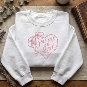May include: A white sweatshirt with a pink heart design. The heart features the text "you are so loved" and the Bible verse John 3:16. A pink bow and ribbons are also part of the design. The sweatshirt is made of a soft material.