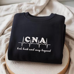 Puede incluir: Una sudadera azul marino con texto bordado en blanco y un diseño de línea de latidos del corazón. El texto dice "CNA" encima de la frase "bed, bath and way beyond". La sudadera está doblada cuidadosamente sobre una superficie neutra.