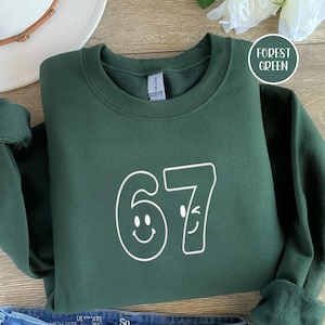 Könnte beinhalten: Forstgrünes Sweatshirt mit der weißen Zahl 67, wobei die Nullen durch Smiley-Gesichter ersetzt werden. Das Sweatshirt ist auf einer Holzoberfläche gefaltet. Ein weißer Hut und eine Blume befinden sich im Hintergrund.