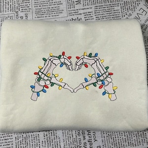May include: Cream-colored sweatshirt featuring an embroidered design of two skeleton hands forming a heart shape. The hands are adorned with colorful Christmas lights, creating a festive and unique design.