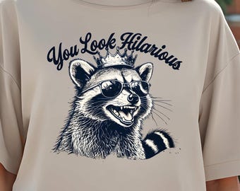 May include: A beige t-shirt with a graphic of a raccoon wearing sunglasses and a crown. The text "You Look Hilarious" is printed above the raccoon.