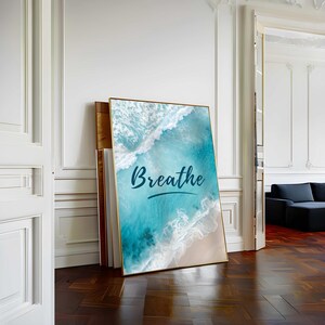 May include: A framed art print featuring a beach scene with turquoise water and white waves. The word "Breathe" is written in a cursive font across the image. The artwork is leaning against a wall in a room with hardwood floors.
