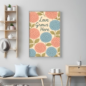 Puede incluir: Arte mural enmarcado con un diseño floral de flores de hortensia melocotón y azul y hojas verdes sobre un fondo crema. El texto "Love Grows Here" está en una fuente cursiva. El marco es plateado.