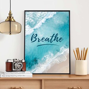May include: A framed art print with the word "Breathe" in cursive over a beach scene with turquoise water and white waves. A vintage camera, books, and pencils in a ceramic holder sit on a wooden table.