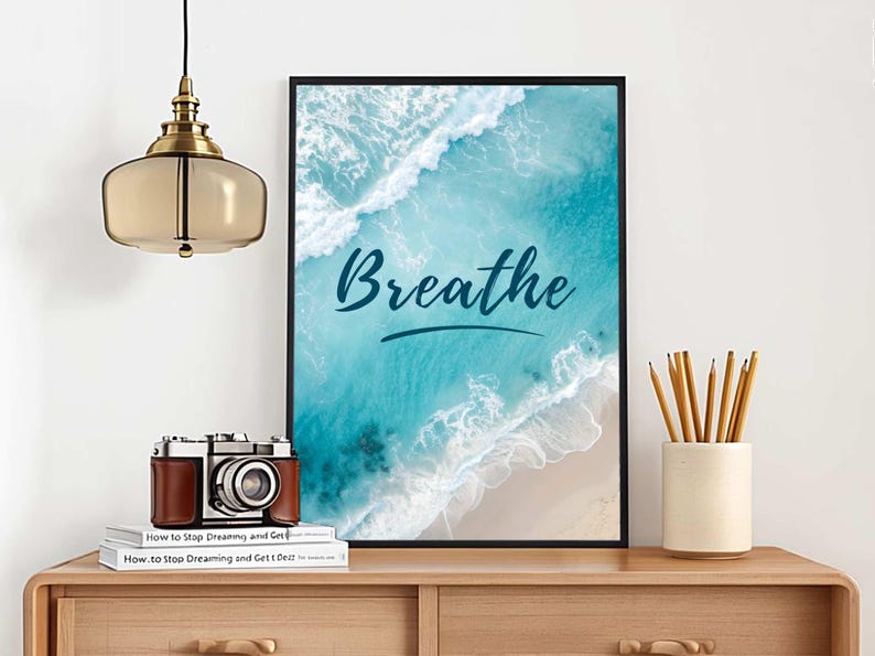May include: A framed art print with the word "Breathe" in cursive over a beach scene with turquoise water and white waves. A vintage camera, books, and pencils in a ceramic holder sit on a wooden table.