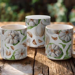 Può includere: Tre tazze in ceramica bianca con un design all'aglio. Le tazze presentano un motivo ripetuto di bulbi d'aglio, spicchi e foglie verdi. Le tazze sono appoggiate su una superficie di legno rustico, con uno sfondo sfocato di verde.
