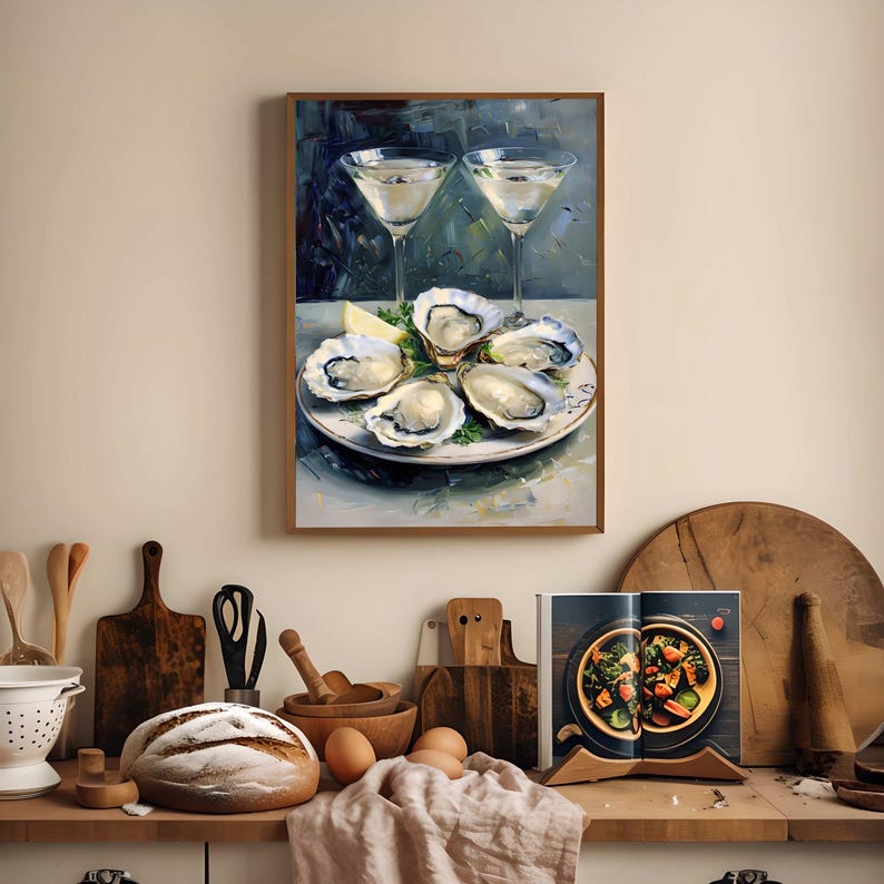 May include: A framed painting depicts two martini glasses and a plate of oysters with lemon. The painting is displayed above a kitchen counter with bread, eggs, and a cookbook. The artwork has a neutral color palette.