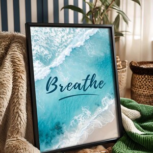 May include: Framed art print featuring a vibrant aerial view of a turquoise ocean with white waves crashing on a sandy beach. The word "Breathe" is written in cursive script across the image. The black frame rests on a cozy blanket.