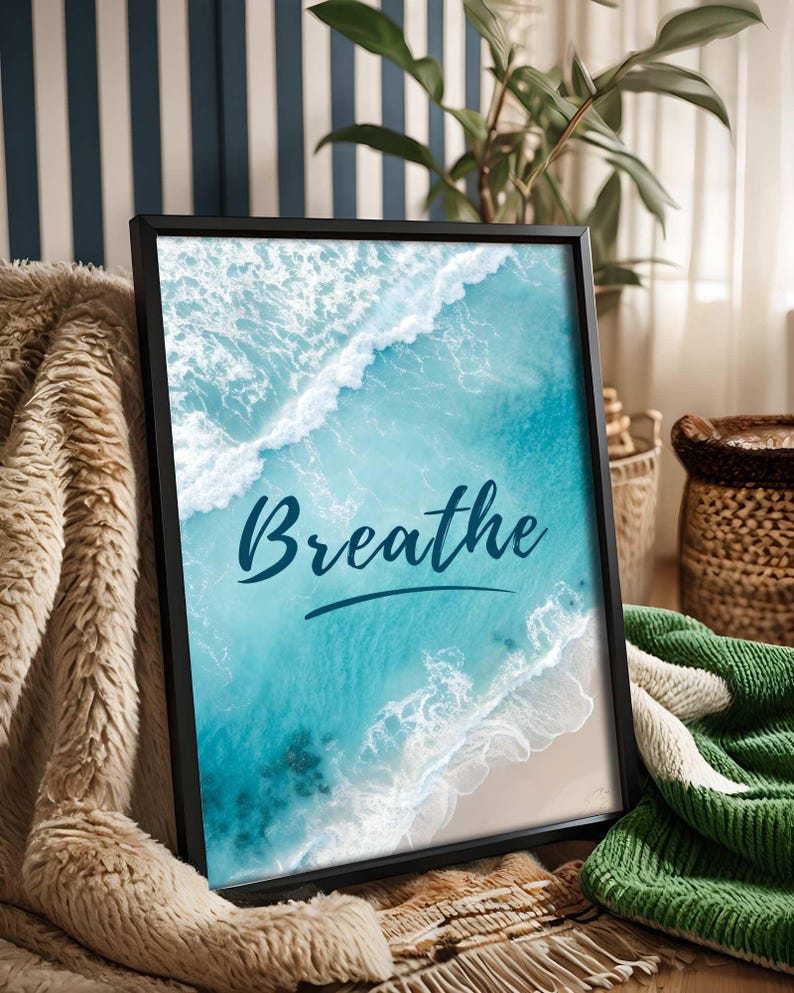 May include: Framed art print featuring a vibrant aerial view of a turquoise ocean with white waves crashing on a sandy beach. The word "Breathe" is written in cursive script across the image. The black frame rests on a cozy blanket.