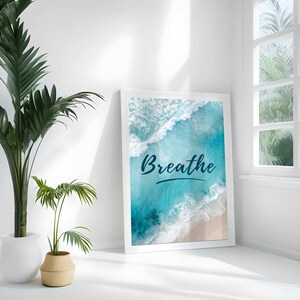 May include: White framed art print featuring an aerial view of ocean waves and a sandy beach. The word "Breathe" is written in a dark blue script across the image. The print is leaning against a white wall, next to a potted plant.