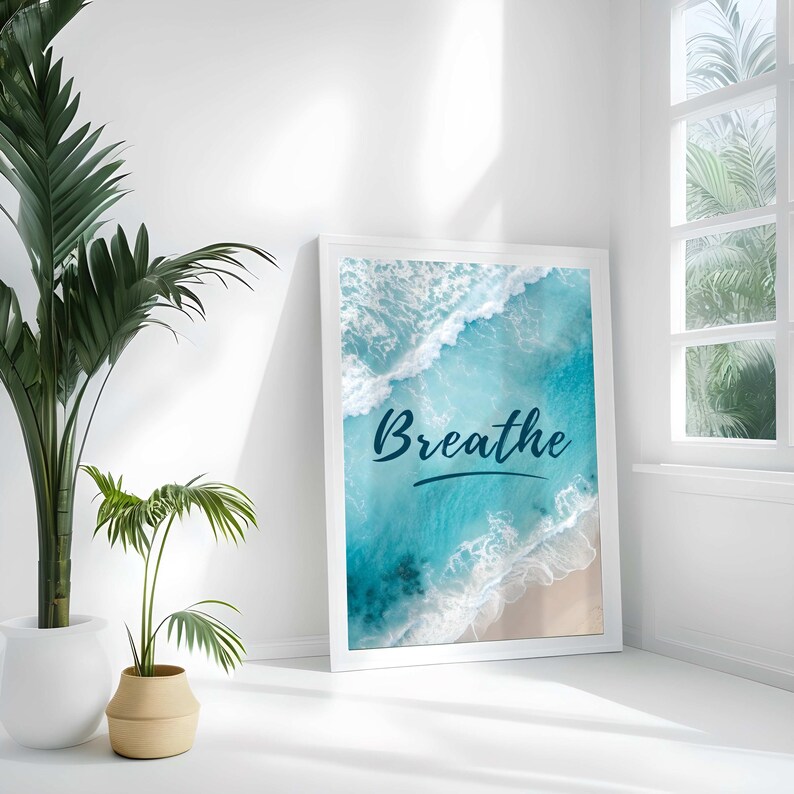 May include: White framed art print featuring an aerial view of ocean waves and a sandy beach. The word "Breathe" is written in a dark blue script across the image. The print is leaning against a white wall, next to a potted plant.