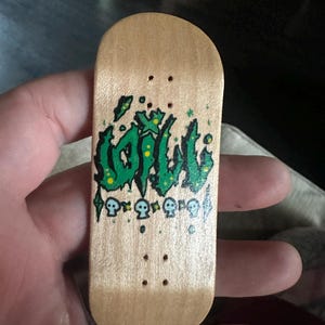 Peut inclure: Une petite planche à doigts en bois avec un motif de style graffiti vert et jaune qui se lit "OIL" et une rangée de têtes de mort. La planche à doigts comporte quatre trous pré-percés pour les trucks.