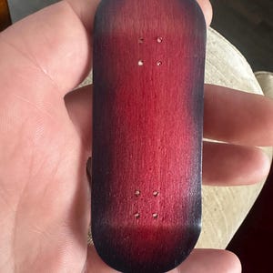 Peut inclure: Une petite planche de fingerboard en bois avec une finition dégradée. La planche passe d'un rouge profond au centre à un bord sombre, presque noir. Quatre petits trous sont percés près du haut et du bas pour le montage des trucks. La planche est tenue dans une main.