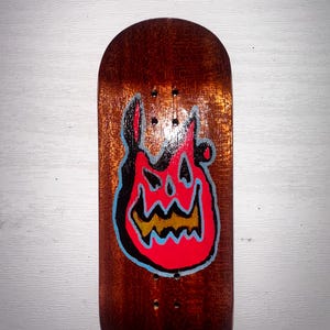 Peut inclure: Une planche à doigts en bois avec un motif de flamme. La planche à doigts est marron avec un motif de flamme rouge, noir et jaune. Elle comporte quatre petits trous pour les trucks.