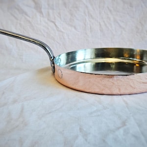May include: A copper-colored frying pan with a silver-toned handle. The pan has a hammered texture on the exterior and a smooth, reflective interior. The handle curves upward, and the pan sits on a white surface.