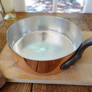 Può includere: Una casseruola con fondo in rame, interno argentato e manico in metallo scuro. La casseruola è appoggiata su un tagliere di legno. Un articolo per la cucina.