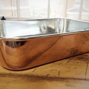 Op de afbeelding: Een koperkleurige rechthoekige braadslede met een glanzend zilveren interieur. De pan heeft afgeronde hoeken en kleine handgrepen aan elke kant. De pan staat op een houten oppervlak.