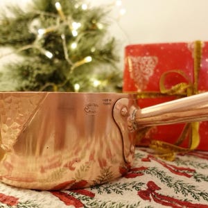 May include: A copper saucepan with a long handle, set against a festive backdrop. The pan has a hammered texture and the text "MADE IN FRANCE". A wrapped gift and a Christmas tree with lights are in the background.