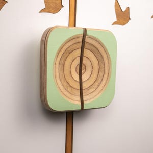 May include: Wooden wall art with a square, pale green frame and a circular, natural wood design. A thin, dark brown band wraps around the frame. The art is mounted on a wooden pole, with wooden bird cutouts in the background.