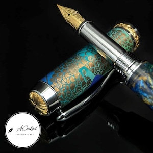 May include: A luxury fountain pen with a gold nib and a detailed, ornate design. The pen features a silver body with a cap adorned with teal and gold patterns. The pen is displayed against a black background.