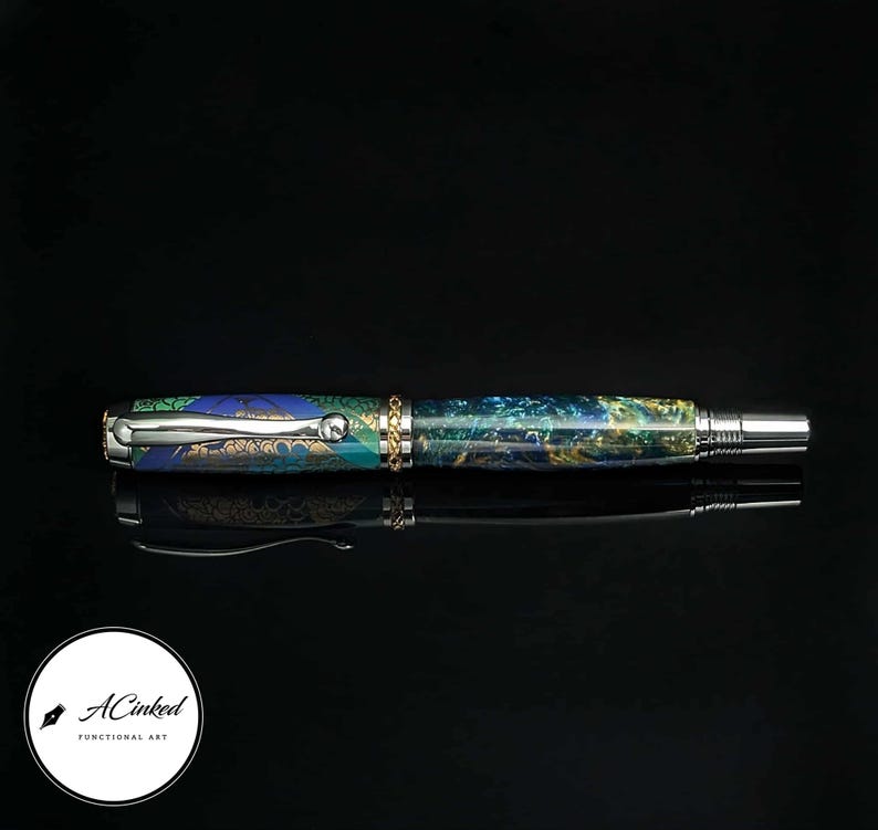 May include: A handcrafted fountain pen with a vibrant, swirling pattern of blues, greens, and gold. The pen features a silver-colored clip and accents, with a gold-colored band near the center. The pen is displayed on a reflective black surface.