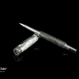 以下が含まれることがあります： シルバーカラーの金属ボディと白黒の大理石模様が特徴の高級ペン。ペンのキャップは外されており、ペン先が見えています。ペンは黒い表面に置かれています。