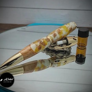 Könnte beinhalten: Ein goldener und brauner Stift mit einem weißen und gelben Marmormuster. Der Stift hat eine goldene Kappe und eine goldene Klammer. Der Stift liegt auf einer reflektierenden Oberfläche. Eine kleine Glasflasche mit schwarzem Verschluss befindet sich ebenfalls auf der Oberfläche.