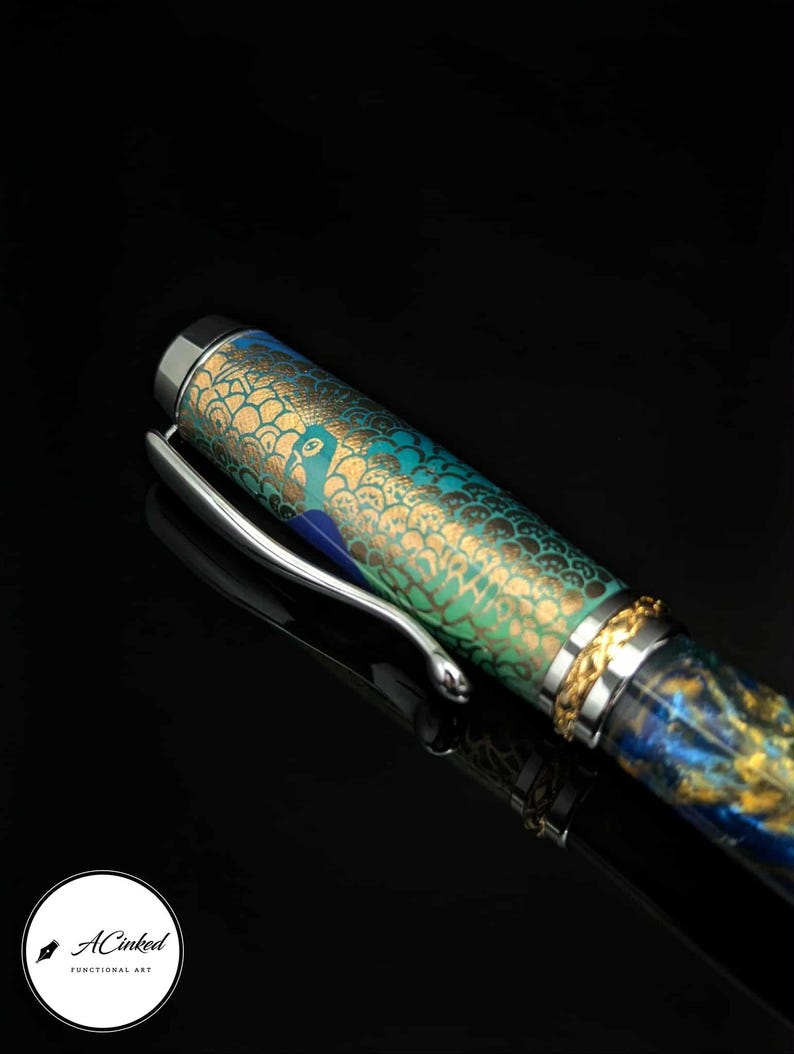 May include: A decorative pen with a peacock design. The pen features a silver clip and a cap with a gold and blue pattern. The barrel of the pen is decorated with a peacock design in shades of green and gold.