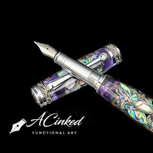 Puede incluir: Dos plumas estilográficas con detalles plateados e incrustaciones iridiscentes de concha de abulón. Las plumas tienen una base de resina púrpura y están sobre un fondo negro. El texto "ACinked FUNCTIONAL ART" se muestra debajo.